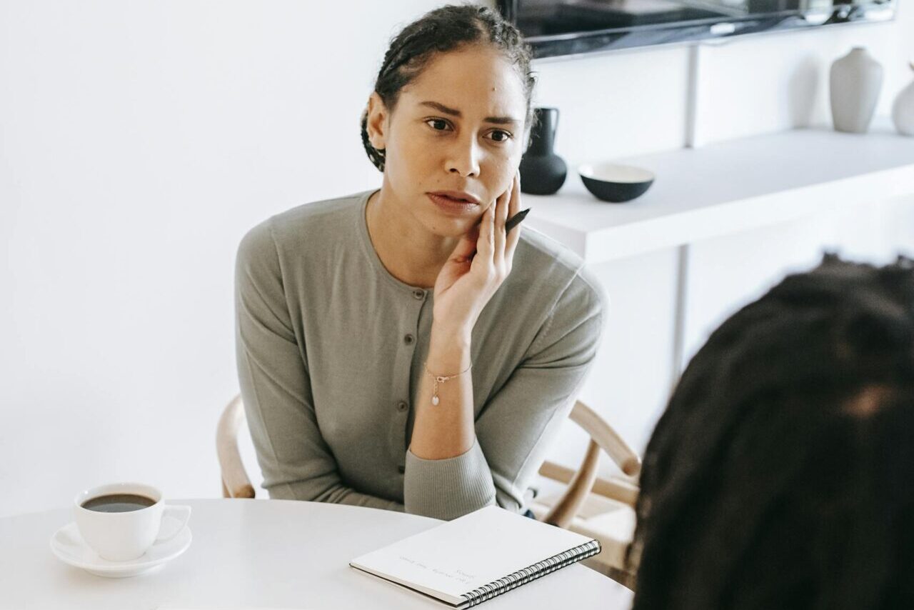 Serious ethnic female consultant listening to black male client problems while sitting at round table together in light psychology center